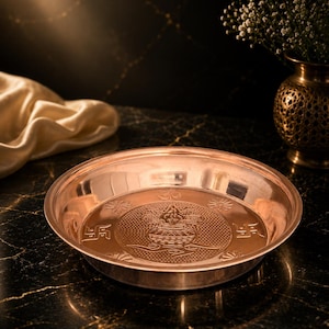 Pode incluir: Um prato redondo de cobre com um design central em relevo de um vaso kalash, rodeado por símbolos de suástica e motivos florais. O prato tem uma superfície metálica polida e reflexiva com uma borda lisa e uma base circular rasa.