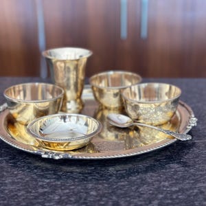 May include: A polished brass dining set consisting of a round serving tray with a beaded edge, a tall tumbler, three small bowls, and a single spoon. The items feature a reflective metallic finish and are arranged together on the circular tray.