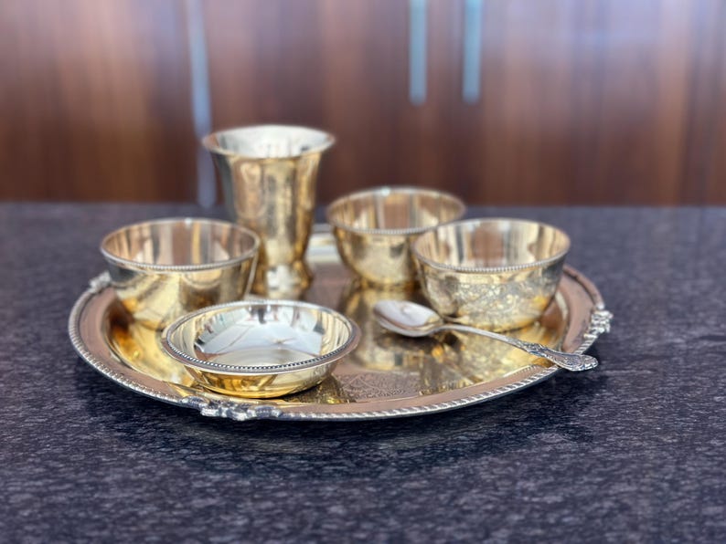 May include: A polished brass dining set consisting of a round serving tray with a beaded edge, a tall tumbler, three small bowls, and a single spoon. The items feature a reflective metallic finish and are arranged together on the circular tray.
