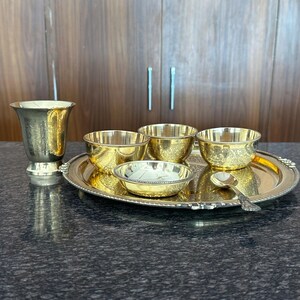 May include: A set of gold-colored metal tableware including a tall flared cup, three small bowls, a shallow dish, and a small spoon, all arranged on a round serving tray with a beaded edge. The items feature intricate engraved floral patterns.