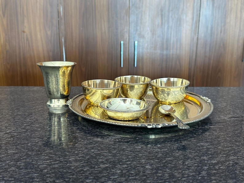 May include: A set of gold-colored metal tableware including a tall flared cup, three small bowls, a shallow dish, and a small spoon, all arranged on a round serving tray with a beaded edge. The items feature intricate engraved floral patterns.