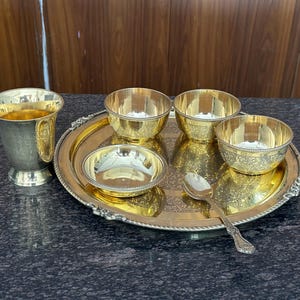 May include: A gold-toned metal serving set consisting of a round tray with an ornate border, three small bowls with textured sides, one shallow bowl, and a single spoon. A tall, flared metal cup with a decorative band sits beside the tray.