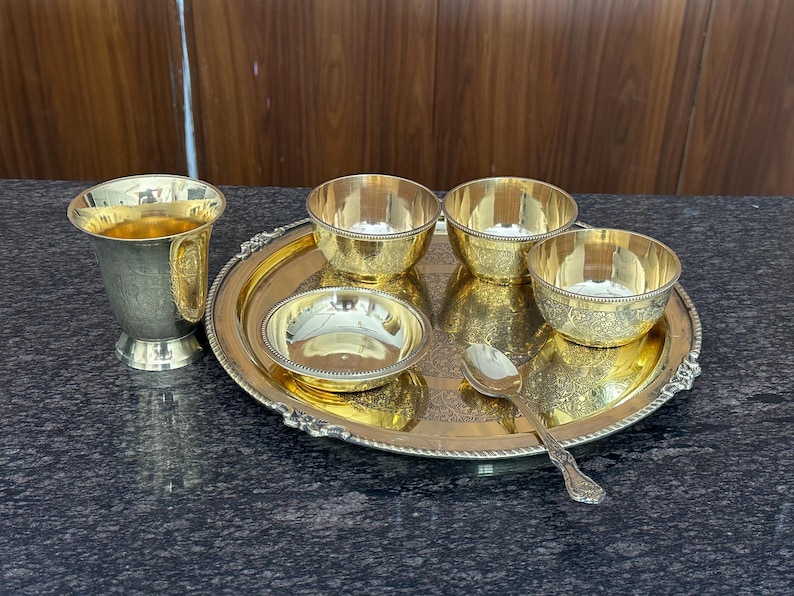 May include: A gold-toned metal serving set consisting of a round tray with an ornate border, three small bowls with textured sides, one shallow bowl, and a single spoon. A tall, flared metal cup with a decorative band sits beside the tray.