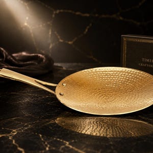 May include: A gold-colored metal pan with a round, hammered texture surface and a long, slender handle attached by two rivets. A black rectangular box is positioned behind the pan with the text "TIMELESS CRAFTSMANSHIP" printed in gold.