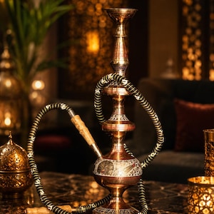 May include: A tall, ornate brass hookah featuring a bulbous base with intricate silver-toned peacock engravings. The central stem is tiered with decorative metalwork, and a long, flexible black hose with white patterns and a light wooden handle is attached.