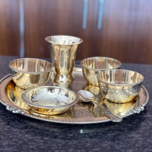 May include: A gold-toned metal serving set consisting of a round tray with a beaded edge, a tall tumbler, three small bowls, and a single spoon. Each piece features intricate, etched floral patterns and a polished, reflective finish.