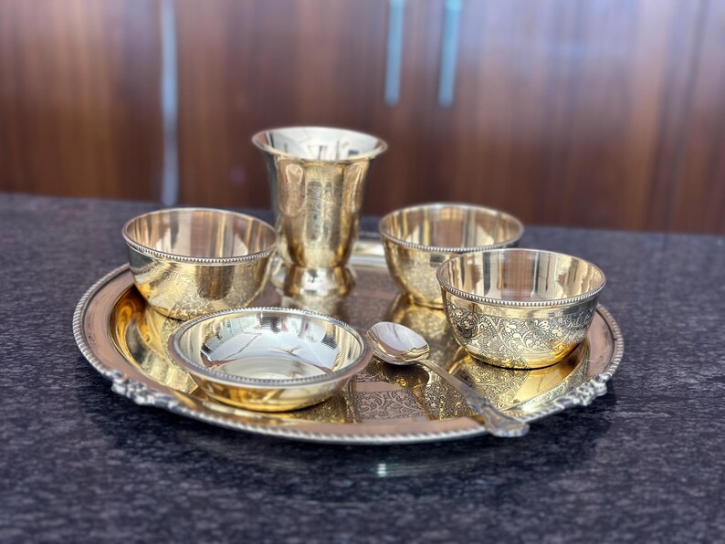 May include: A gold-toned metal serving set consisting of a round tray with a beaded edge, a tall tumbler, three small bowls, and a single spoon. Each piece features intricate, etched floral patterns and a polished, reflective finish.