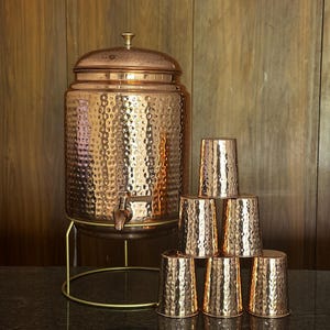 May include: A copper water dispenser with a hammered texture, a spigot, and a lid, set on a gold-colored stand. Six matching copper cups are stacked nearby. The dispenser and cups have a reflective surface.