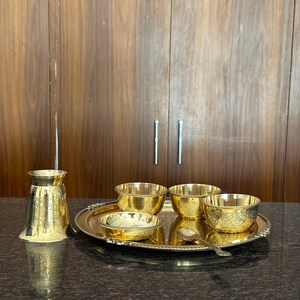 May include: A polished brass serving set consisting of a round tray, three small bowls, a shallow dish, a tall tumbler, and a small spoon. Each piece features intricate etched floral patterns and a bright, reflective gold-toned metallic finish.