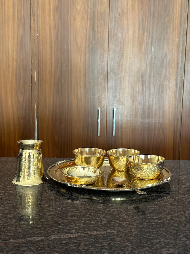 May include: A polished brass serving set consisting of a round tray, three small bowls, a shallow dish, a tall tumbler, and a small spoon. Each piece features intricate etched floral patterns and a bright, reflective gold-toned metallic finish.