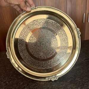 May include: A round brass serving tray featuring an intricate engraved floral pattern in the center and a decorative scalloped edge with embossed detailing. The metallic surface has a polished finish with a warm golden tone and a smooth rim.