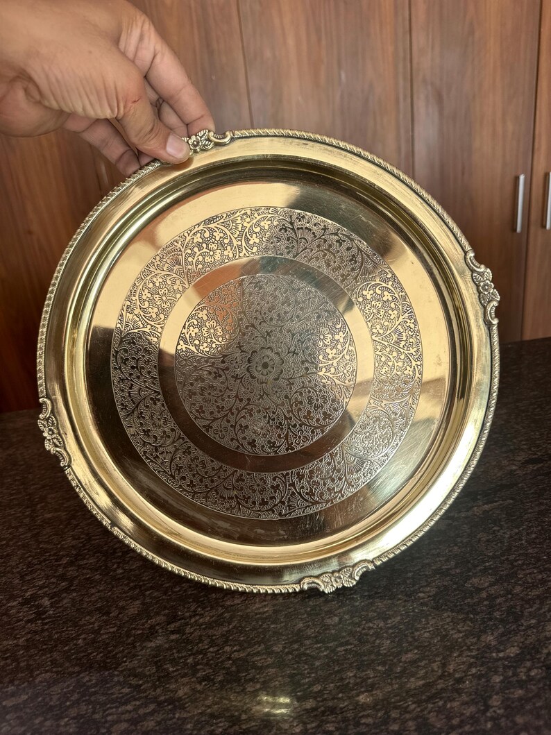 May include: A round brass serving tray featuring an intricate engraved floral pattern in the center and a decorative scalloped edge with embossed detailing. The metallic surface has a polished finish with a warm golden tone and a smooth rim.