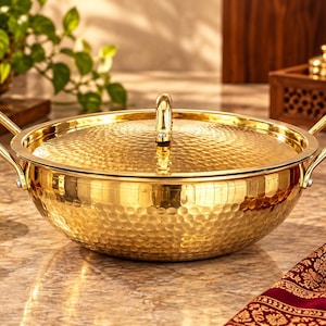 May include: A round, gold-colored metal cooking pot with a hammered texture, featuring two side handles and a matching lid with a central handle. The polished surface reflects light, highlighting the dimpled pattern across the exterior.