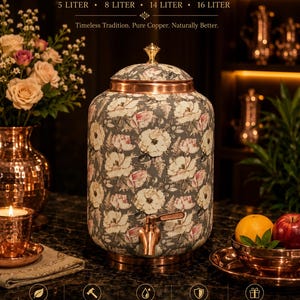 May include: A cylindrical copper water pot featuring a dark gray exterior with a white and pink floral pattern. The pot includes a metallic copper lid with a small knob, a copper spigot at the base, and a copper bottom rim. Text reads COPPER WATER POT.