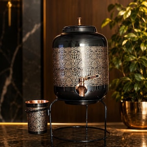 May include: A black beverage dispenser with a metallic gold damask pattern and a copper-toned spigot labeled SAGA. The dispenser sits on a black metal stand with an attached matching cup holder containing a black and gold cup.