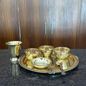 May include: A polished brass dining set consisting of a round serving tray with a beaded edge, three small bowls with intricate floral engravings, a tall tapered drinking cup, and a single matching spoon, all featuring a bright metallic finish.