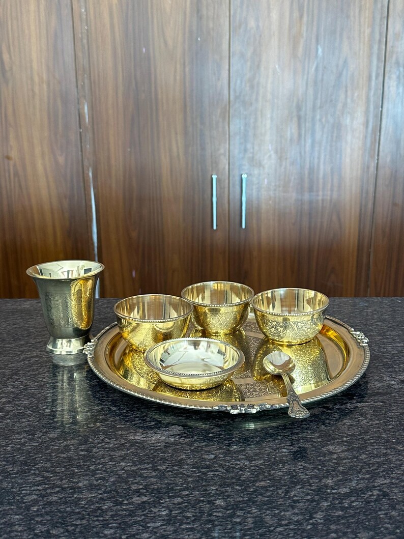 May include: A polished brass dining set consisting of a round serving tray with a beaded edge, three small bowls with intricate floral engravings, a tall tapered drinking cup, and a single matching spoon, all featuring a bright metallic finish.