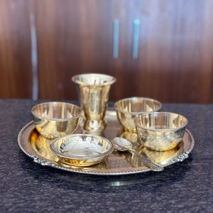 May include: A polished brass dining set consisting of a large round serving tray with a beaded edge, three small bowls with intricate floral engravings, one tall tapered tumbler, and a single matching spoon, all arranged on a dark surface.