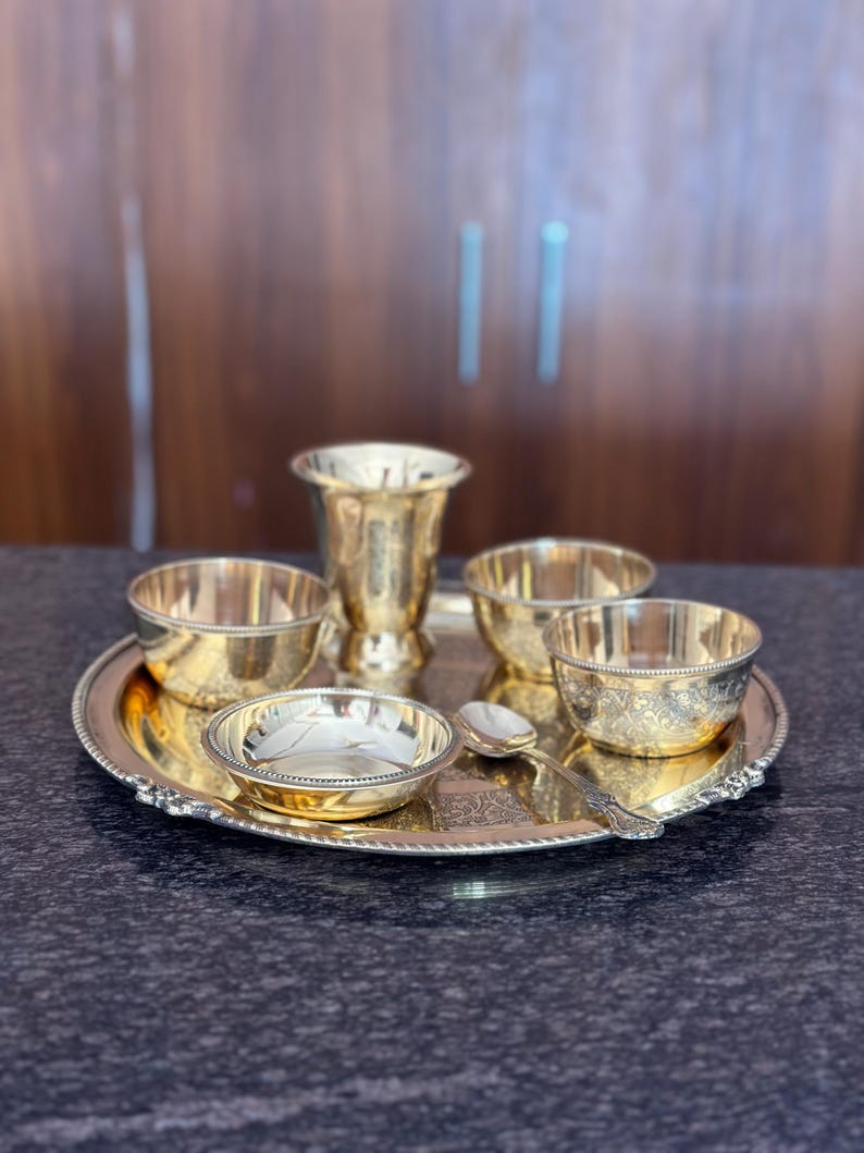 May include: A polished brass dining set consisting of a large round serving tray with a beaded edge, three small bowls with intricate floral engravings, one tall tapered tumbler, and a single matching spoon, all arranged on a dark surface.