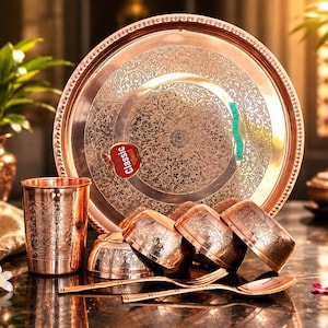 May include: A copper-coloured metal dinnerware set featuring a large round tray with intricate floral engravings, a matching tumbler, three small bowls, a fork, and a spoon. A red apple-shaped sticker with the text Classic is on the tray.