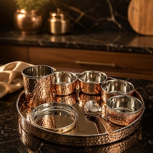 May include: A round serving tray with a hammered copper exterior and a smooth metallic interior, containing a tall hammered copper tumbler, four small hammered copper bowls, a small flat plate, and a single polished metal spoon.