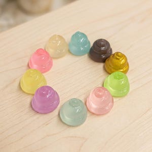 May include: A collection of ten small, smooth, swirl-shaped resin charms arranged in a circle. The charms feature a variety of translucent and opaque colors, including pink, yellow, purple, light green, blue, brown, and a metallic gold finish.