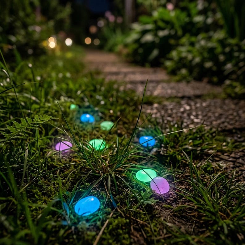 May include: A collection of small, smooth, oval-shaped decorative stones scattered throughout green grass. These stones emit a soft, glowing light in various colors, including blue, green, and purple, creating a luminous effect in the dark.