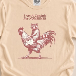Puede incluir: Una camiseta de manga corta de color beige claro con un gráfico granate de un mapache montado sobre un gallo. Sobre la ilustración, el texto dice, I Am A Conduit For NONSENSE. La camiseta tiene cuello redondo y una etiqueta de la marca Gildan.