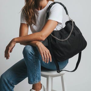 May include: A black nylon hobo bag featuring a bamboo-style top handle, a silver-toned metal chain accent across the front, and a long adjustable shoulder strap. The bag has a soft, slouchy silhouette with a secure zipper closure at the top.