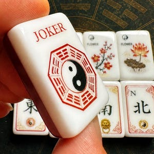 May include: A white Mahjong tile featuring the word JOKER in red text above a red and black yin-yang symbol surrounded by an octagonal trigram design. Additional Mahjong tiles with floral, directional, and symbolic graphics are visible in the background.