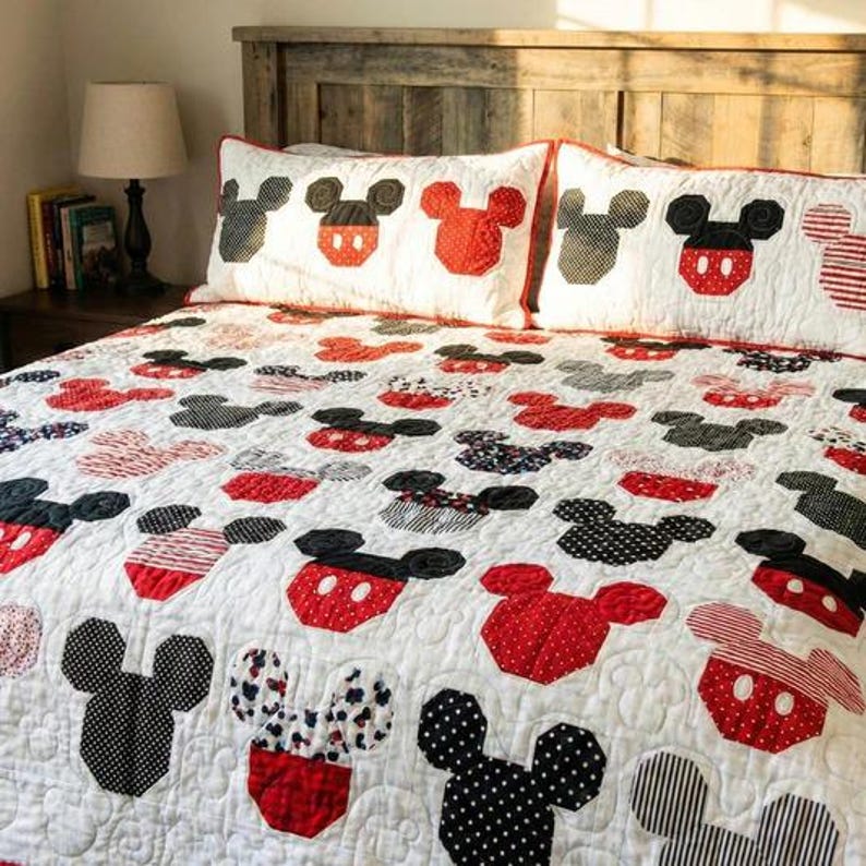 May include: A white quilt and two pillowcases feature a repeating pattern of Mickey Mouse heads in red, black, and patterned fabrics. The quilt and pillowcases are on a wooden bed frame.
