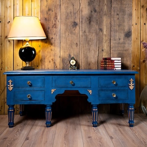 Peut inclure: Un bureau bleu avec des détails dorés et six tiroirs. Une lampe noire avec un abat-jour crème est posée sur le bureau, ainsi qu'une petite horloge et des livres. Un grand vase en verre avec des fleurs séchées est à droite.