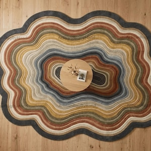 Può includere: Un tappeto dalla forma irregolare caratterizzato da fasce concentriche e ondulate di colore grigio scuro, terracotta, giallo senape, verde salvia e crema. Il tappeto presenta un aspetto strutturato e stratificato, circondando un tavolino rotondo in legno.