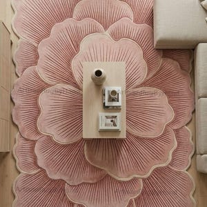 May include: A large, soft pink area rug shaped like a blooming flower with layered, scalloped petals featuring fine, radiating line textures. The rug has a light beige border and the text ArtisanRugByHand is visible in multiple locations.