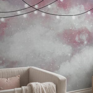Puede incluir: Un mural de pared con un cielo estilo acuarela con nubes suaves en rosa y gris, acentuado por motas blancas y una cadena de luces brillantes colgada en la parte superior. Una cuna beige con una almohada rosa está en primer plano.