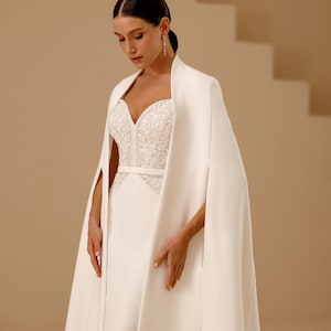 May include: A floor-length white gown featuring a sweetheart neckline with intricate silver beadwork and embroidery on the bodice, paired with a matching floor-length cape that drapes over the shoulders with open arm slits and a smooth finish.