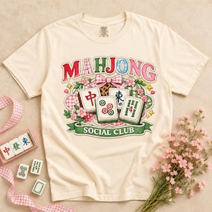 Puede incluir: Una camiseta de manga corta de color crema con un estampado gráfico que incluye el texto "MAHJONG SOCIAL CLUB". El diseño presenta ilustraciones de cuatro fichas de mahjong, flores rosas, un lazo a cuadros y pancartas decorativas verdes.