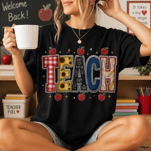 Pode incluir: Uma t-shirt preta de manga curta com a palavra TEACH em letras grandes e estampadas. O T é de xadrez vermelho, o E tem girassóis, o A é preto com ícones escolares, o C é de ganga azul e o H tem lápis e clipes desenhados.