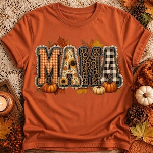 Peut inclure: Un t-shirt à manches courtes orange brûlé avec le mot MAMA en grandes lettres majuscules. Chaque lettre présente un motif unique incluant pied-de-poule, tournesols, imprimé léopard et carreaux vichy, agrémenté de feuilles d'automne et de citrouilles.