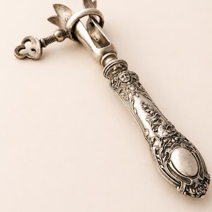 May include: An antique silver wine bottle opener with ornate floral and face detailing. The opener features a key-shaped handle and a decorative design on the handle. The opener is made of silver metal.