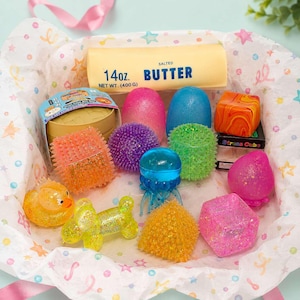 Op de afbeelding: Een verzameling sensorisch speelgoed, waaronder een boterblok van 400g, een gele eend, een ballonhond, een blauwe kwal en diverse getextureerde blokjes en bollen in oranje, paars, groen, roze en geel, liggend op vloeipapier met sterrenpatroon.