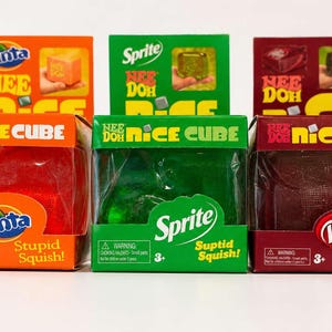 Puede incluir: Tres juguetes Nee Doh Nice Cube en cajas. Las cajas son naranjas, verdes y granates, con los logotipos de Fanta, Sprite y Dr. Pepper. Cada caja tiene una ventana transparente que muestra el cubo en su interior. El texto "Stupid Squish!" está en las cajas.