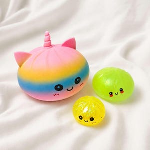 May include: A large rainbow-colored squishy toy shaped like a bun with a unicorn horn and ears, featuring a smiling face. Beside it are two smaller, round squishy toys: one translucent lime green and one yellow with glitter, both with smiling faces.