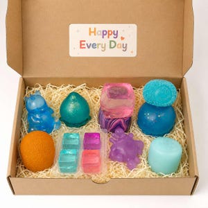 May include: A cardboard gift box containing various colorful soap shapes, including a blue bear, a teal peach, a pink cube, a blue smiley face sphere, an orange sphere, a light blue cylinder, a purple creature, and four small square soaps with the text Happy Every Day.