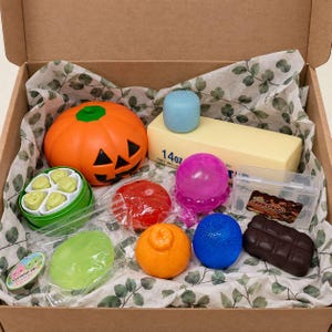 May include: A cardboard box containing a collection of items including an orange jack-o'-lantern, a 397g block of butter with a blue cube on top, a green container with small green shapes, a red sphere, a purple sphere, a blue sphere, and a chocolate bar.