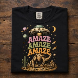 May include: A black t-shirt featuring a graphic print of a flying saucer hovering over a rocky desert landscape with a cactus. The text "AMAZE" is printed three times in stacked, wavy, colourful letters. The design includes planets, stars, and a crescent moon.