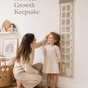 May include: A vertical fabric growth chart featuring a black line drawing of a tall house with multiple windows and a door. The chart includes numerical height markings in centimeters. The text A Sweet Growth Keepsake is printed on the wall.
