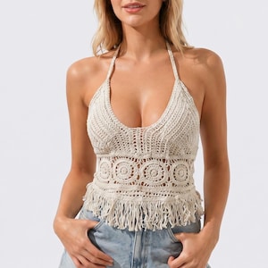 May include: A beige crochet halter top featuring a deep V-neckline, intricate circular patterns across the midsection, and a fringed hemline. The garment is paired with light blue denim shorts that have a frayed, distressed hem detail.