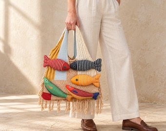 Peut inclure: Un sac fourre-tout en tricot beige avec un panneau bleu clair et cinq appliqués de poissons en relief en forme de peluche, aux motifs rouges, bleu foncé, orange, verts et multicolores. Le sac est orné de longues franges nouées en bas.