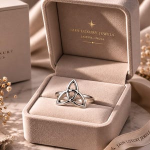 May include: A silver-toned ring featuring a triquetra knot design, presented inside a beige velvet-lined jewelry box. The box lid interior displays the gold-printed text JAIN LUXURY JEWELS JAIPUR, INDIA with a star icon above the brand name.